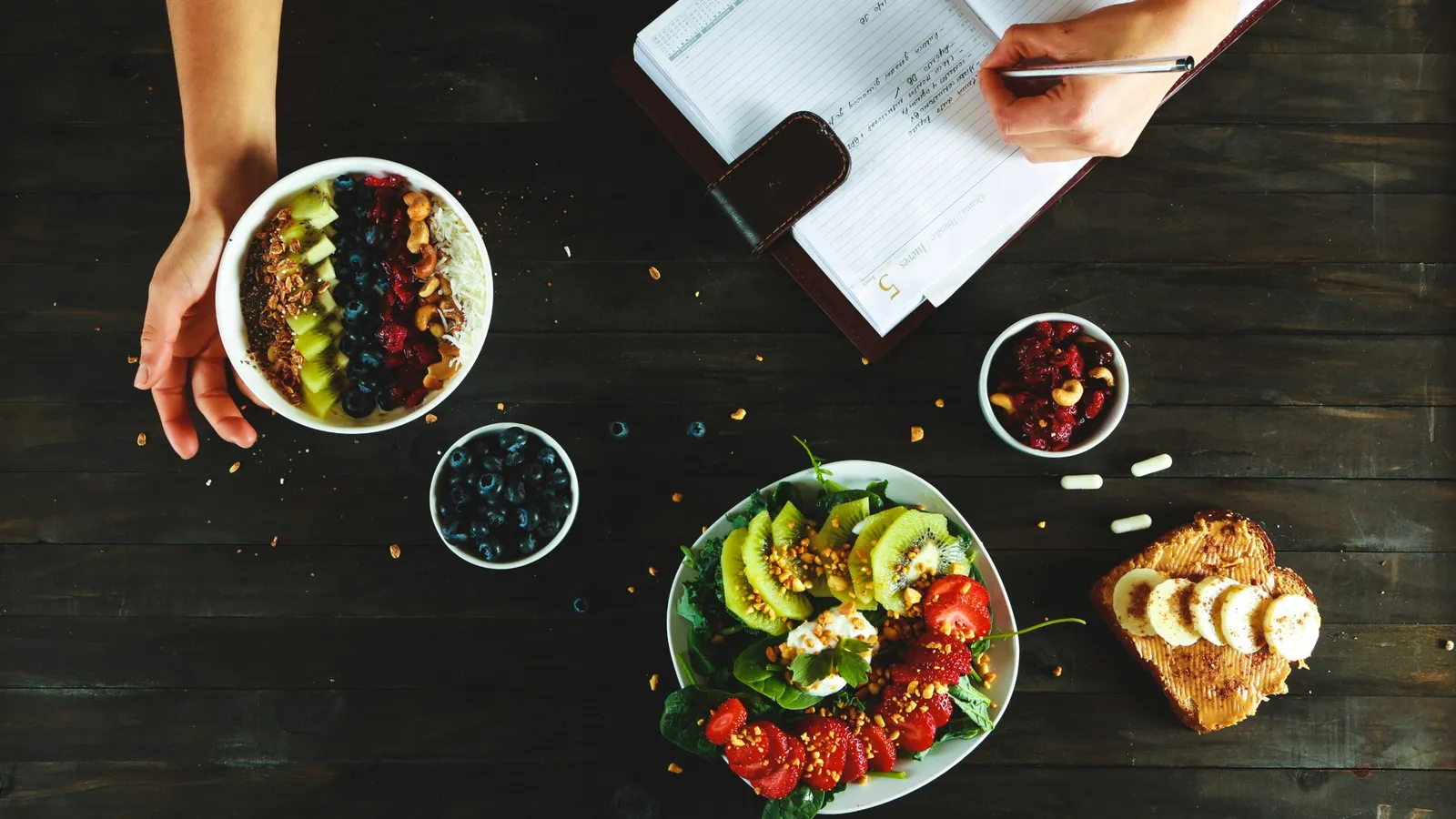 Overhead view of a planner being written beside bowls of fruit, a salad and a few capsules