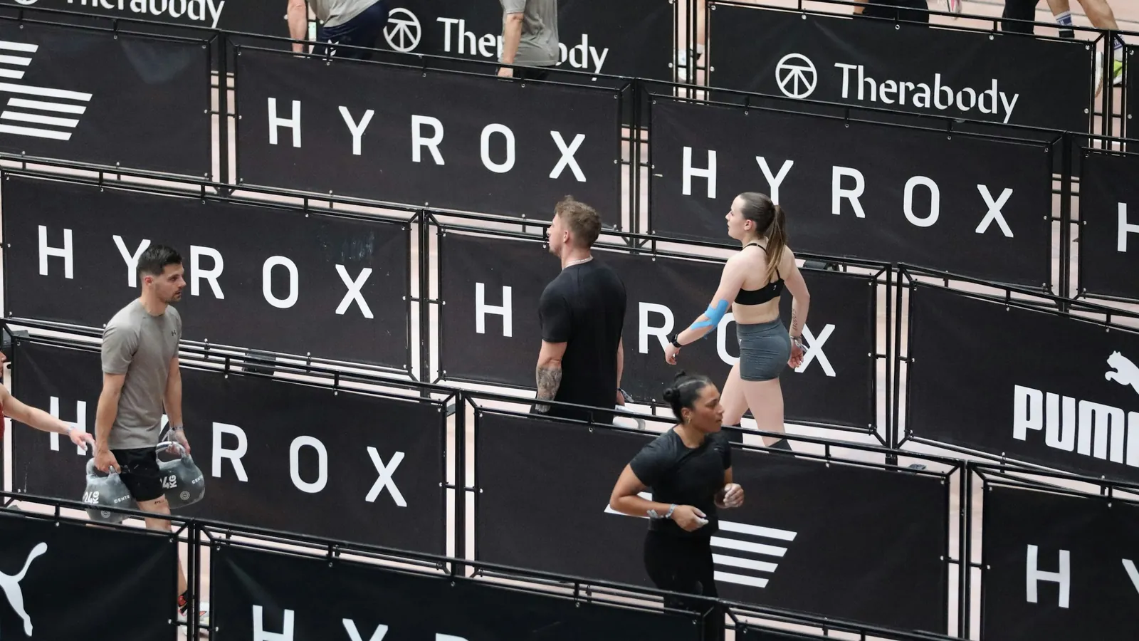Athletes moving between stations at an indoor Hyrox fitness race, the strength and endurance format pulling general clients towards hybrid training