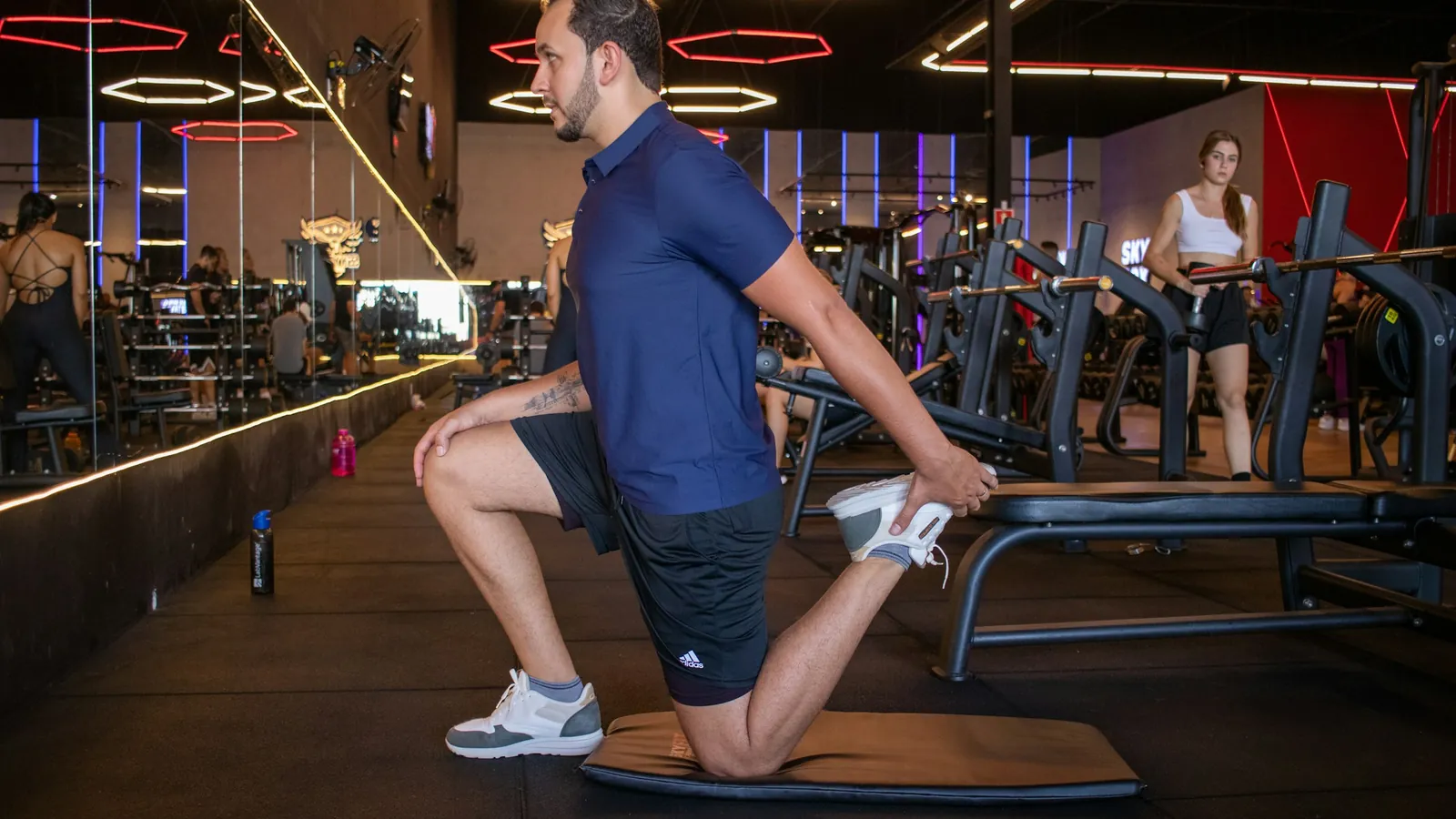 A man in a gym holding a kneeling quad stretch on a mat before training, the warm-up work a coach programmes